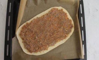 Bekleed een bakplaat met bakpapier en plaats er een platte cake van gistdeeg op.We halen het gehakt uit de koelkast en verdelen de vulling gelijkmatig over het hele oppervlak van het deeg, waarbij er wat ruimte rond de randen overblijft.