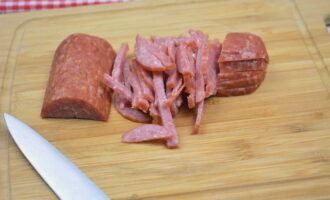 Ang salad ng Hunter na may pinausukang sausage ay napakadaling ihanda. Kumuha ng isang tinapay ng sausage at putulin ang kinakailangang halaga gamit ang isang matalim na kutsilyo. Naglalabas kami ng isang piraso ng sangkap mula sa shell. Ilagay ang sausage sa isang cutting board at hiwain ng manipis na piraso.