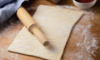 Habang lumalamig ang pagpuno ng pie, gawin natin ang kuwarta. Pre-defrost ito at igulong ang unang layer. Ang hugis ng layer ay hugis-parihaba, ang kapal ay 2-3 millimeters. Kasama ang malawak na gilid hinahati namin ang layer sa kalahati (biswal). Gumagawa kami ng mga pagbawas tuwing 3-4 sentimetro sa isang kalahati, at inilalagay ang pagpuno sa isa pa. Iwanan ang gilid ng kuwarta upang ma-secure ang mga gilid ng mga gilid. 