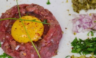 Laten we beginnen met serveren: leg het rundvlees neer zoals op de foto, maak een kleine kuiltje in het midden en leg de dooier neer. Plaats de fijngehakte additieven in de buurt.
