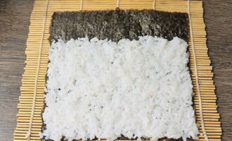 Helyezzen egy nori lapot a szőnyegre, sima oldalával lefelé, vizes ujjaival terítse szét a rizst úgy, hogy ne hagyjon réseket, az egyik oldalon két centimétert kilépve, és ne érje el az ellenkező oldalt. 