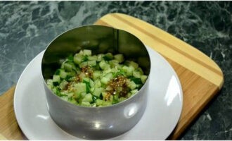 We verzamelen de salade in lagen. Maar je kunt ook gewoon alle gehakte producten in een kom mengen. Plaats een serveerring op een platte schaal. Leg de komkommers in de eerste laag en giet de dressing erover.
